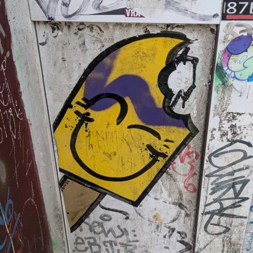 yellow popsicle with black smiley face and black outline cleanly spray painted on gray wall. Surrounded by some tags and stickers