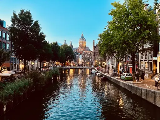 A scenic evening view of a canal in Amsterdam. The canal is flanked by rows of trees and traditional buildings, many of which have warm lights glowing from windows and street lamps. Reflections of these lights shimmer on the calm water. Several boats are moored along the canal’s edge. People are visible strolling or relaxing at outdoor cafes. In the distance, a large church with a dome and spires rises above the rooftops. The sky is clear, fading from blue into the soft hues of dusk.