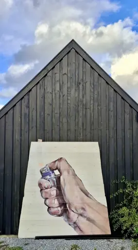 Upright image, a large painting (perhaps 2 metres by 2 metres, not sure) propped up at an angle against a black wooden hut. The hut facade is triangular with a sharp point up into a blue sky with white clouds. The painting's 'canvas' is wooden planks painted cream. Filling it, the image of a white person's hand holding a cheap disposable cigarette lighter. They've clicked it, flame is coming out. I realised how badly I think the world is going when I immediately assumed this was a call to just burn everything down and start the world again. The painting, which presumably had been put outside to dry or was awaiting collection, is by artist Eske Touborg. This is the back of his studio - a wooden hut at the marina at Marselisborg, Aarhus, Denmark.